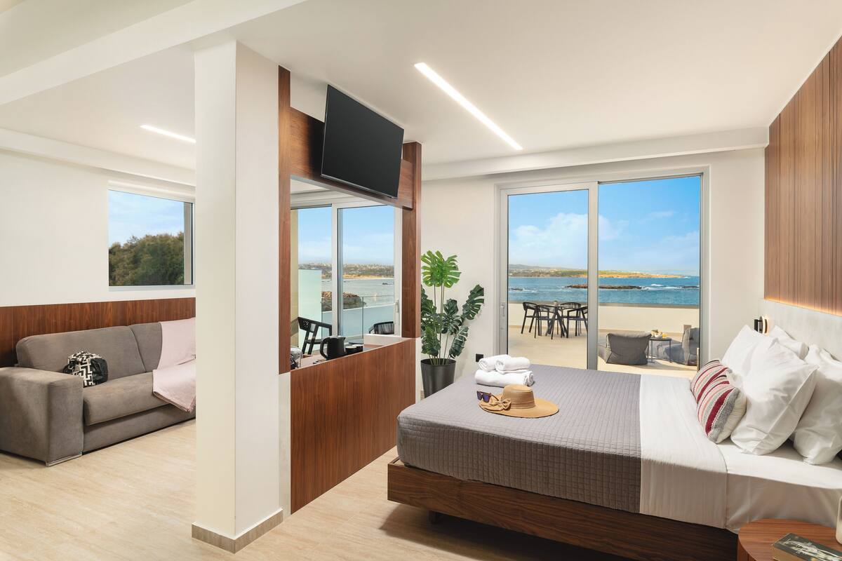 A modern hotel room with a large bed, a sofa, a TV mounted on the wall, and a balcony with a view of the sea.
