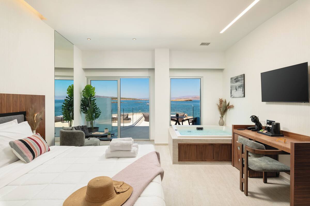 A modern hotel room with a large bed, a desk with a laptop, a view of the sea, and a jacuzzi.