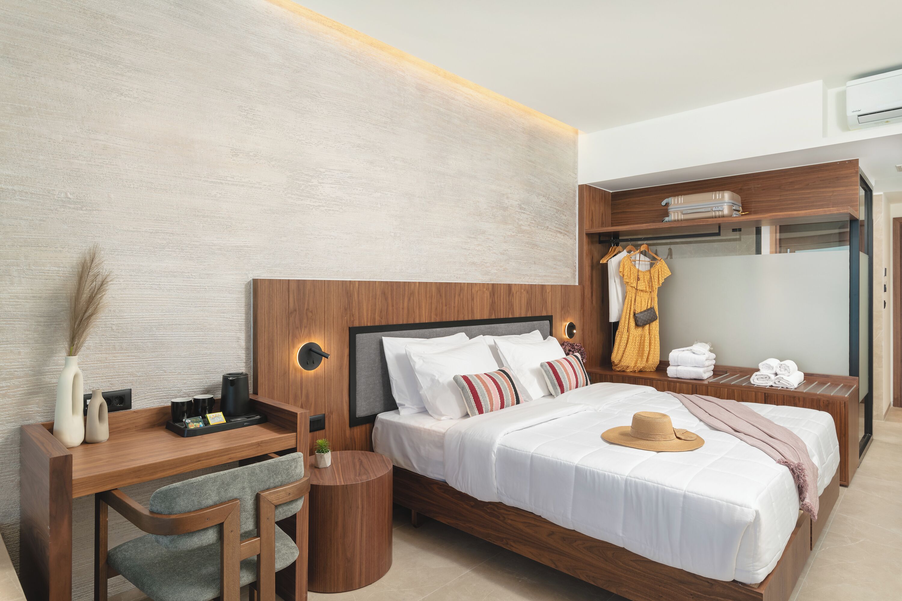A modern hotel room with a wooden bed, a desk with a chair, a vanity with a mirror, and a wardrobe.