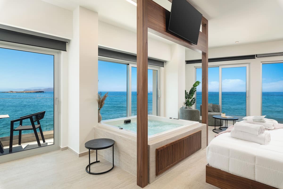 A modern room with a large bathtub, a bed, a TV, and a view of the sea.