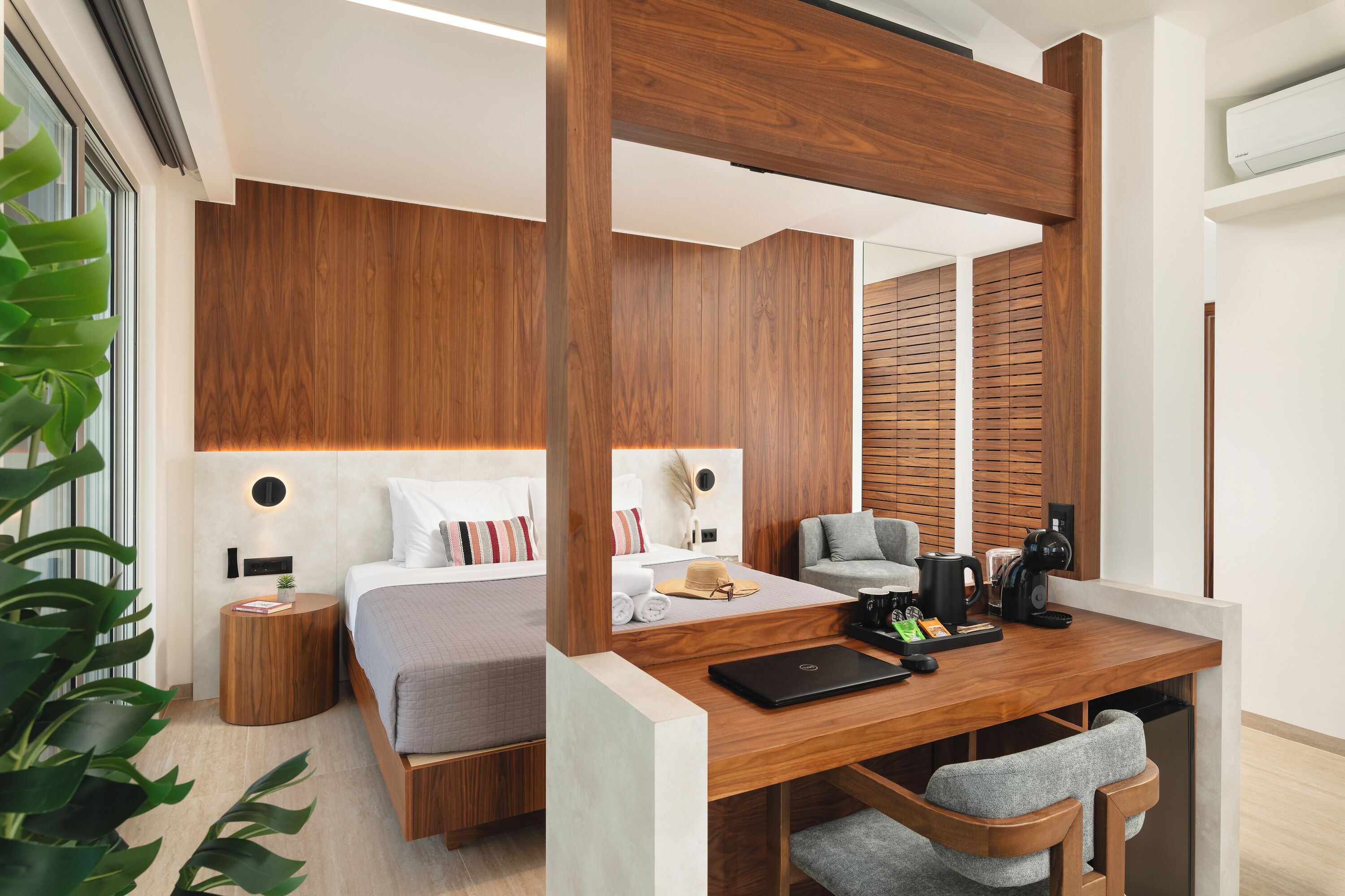 A modern hotel room with a wooden headboard, a bed with white linens, a desk with a laptop, and a seating area with a chair.