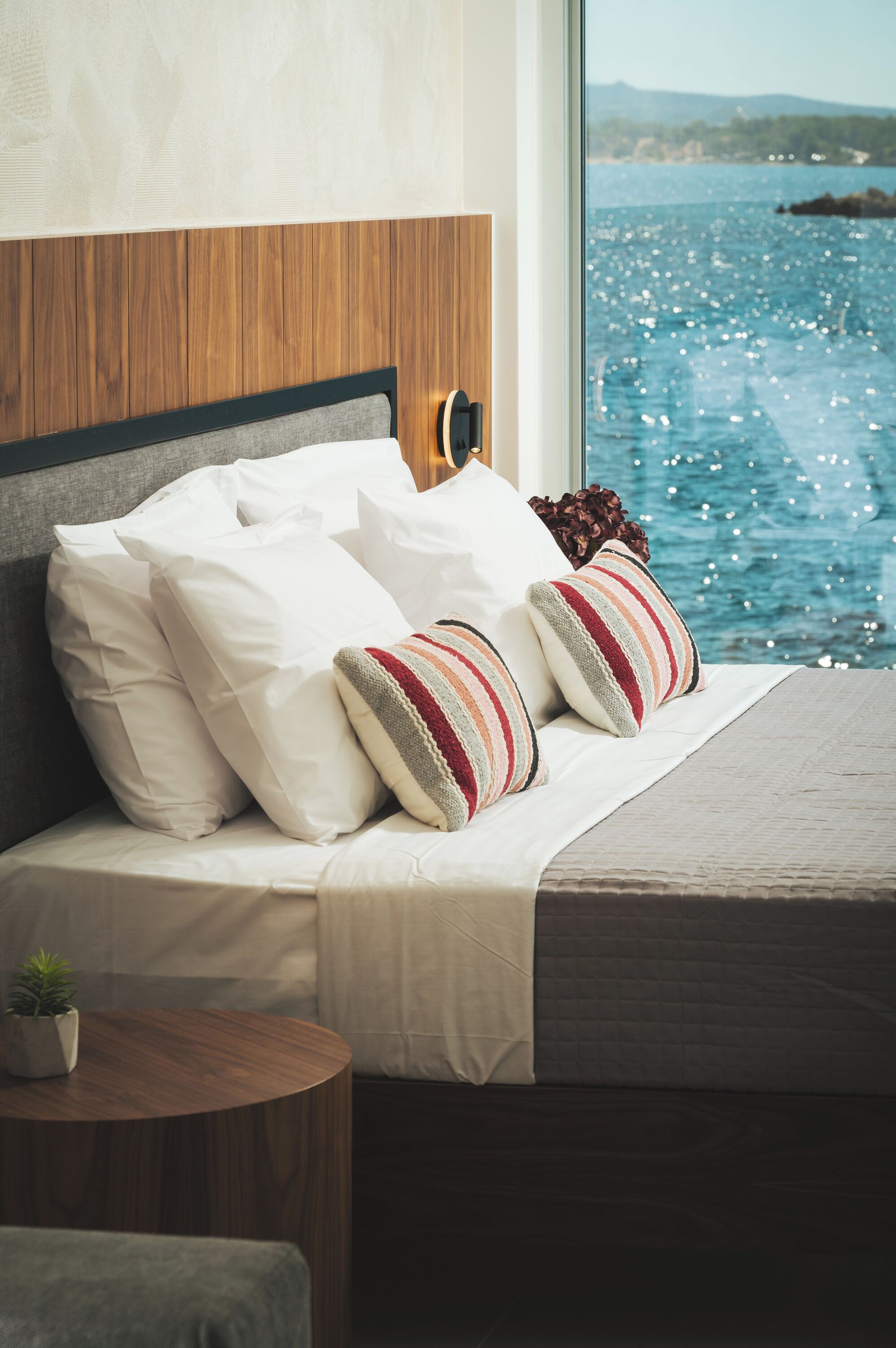 A bed with white and striped pillows, a small round table with a plant, and a view of the sea through a window.