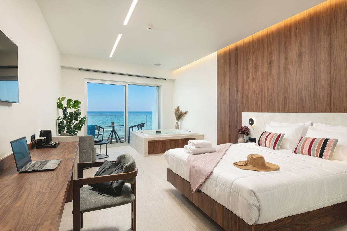 A modern hotel room with a large bed, a desk with a laptop, a view of the ocean, and a balcony.