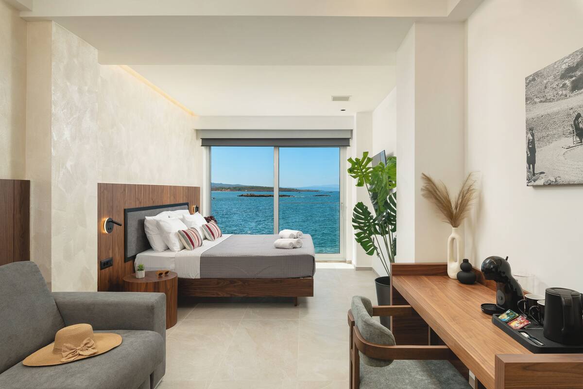 A modern bedroom with a large window, a bed, a desk, and a view of the sea.