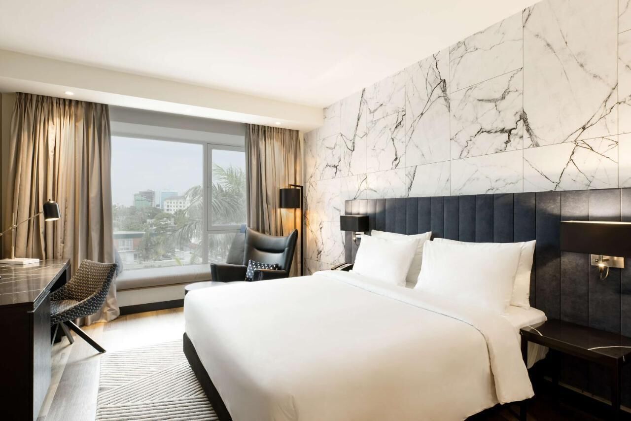 Habitación de hotel con una cama grande, un escritorio, una silla, un revestimiento de pared con diseño de mármol y vistas a la ciudad.