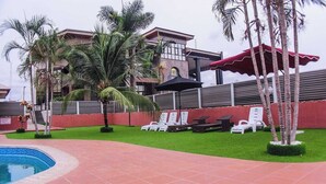 A pool area with lounge chairs, palm trees, and a building with a red wall and a black awning. - De Castle hotels & Resort Enugu (Enugu)