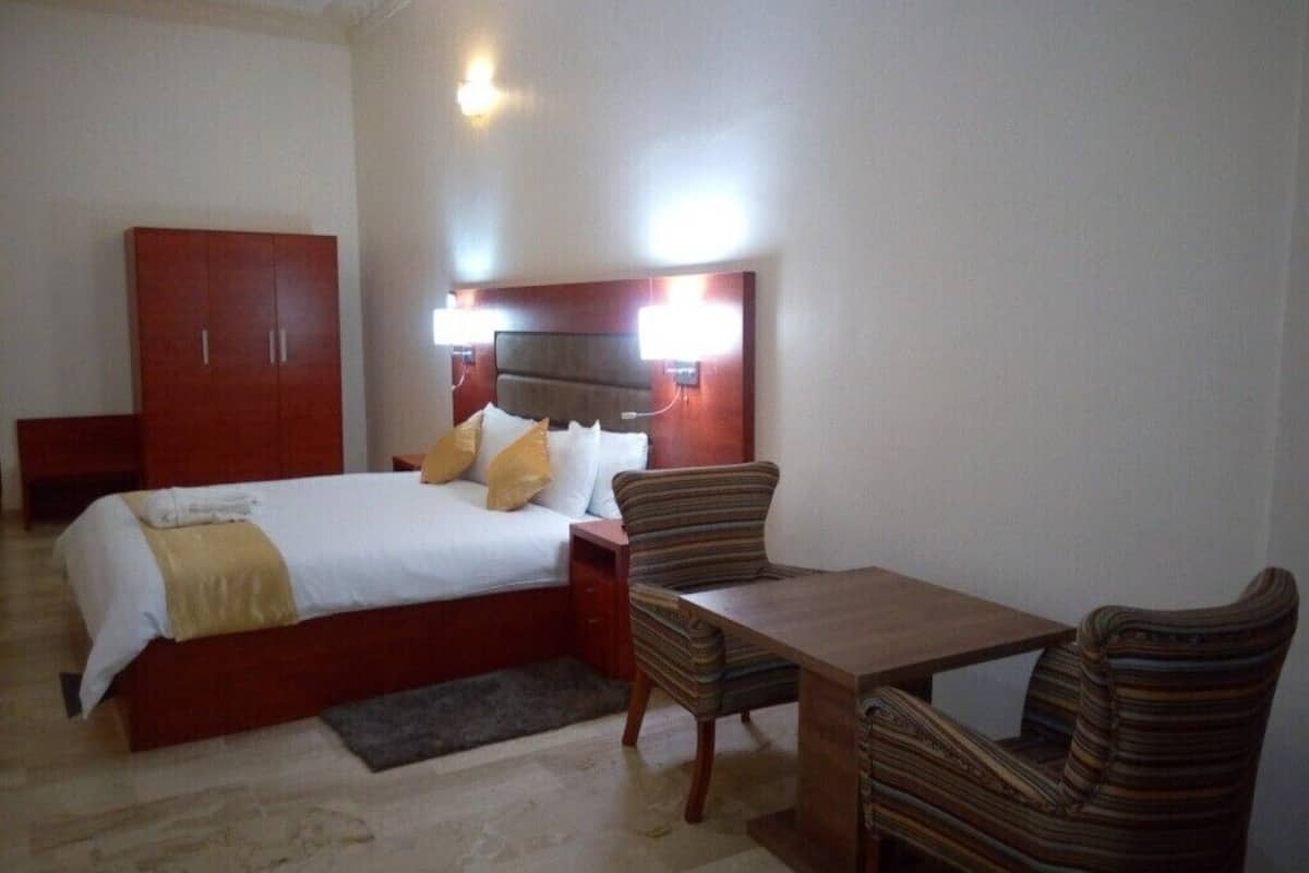 A hotel room with a bed, a wooden nightstand, a striped armchair, a wooden table, and a wardrobe.