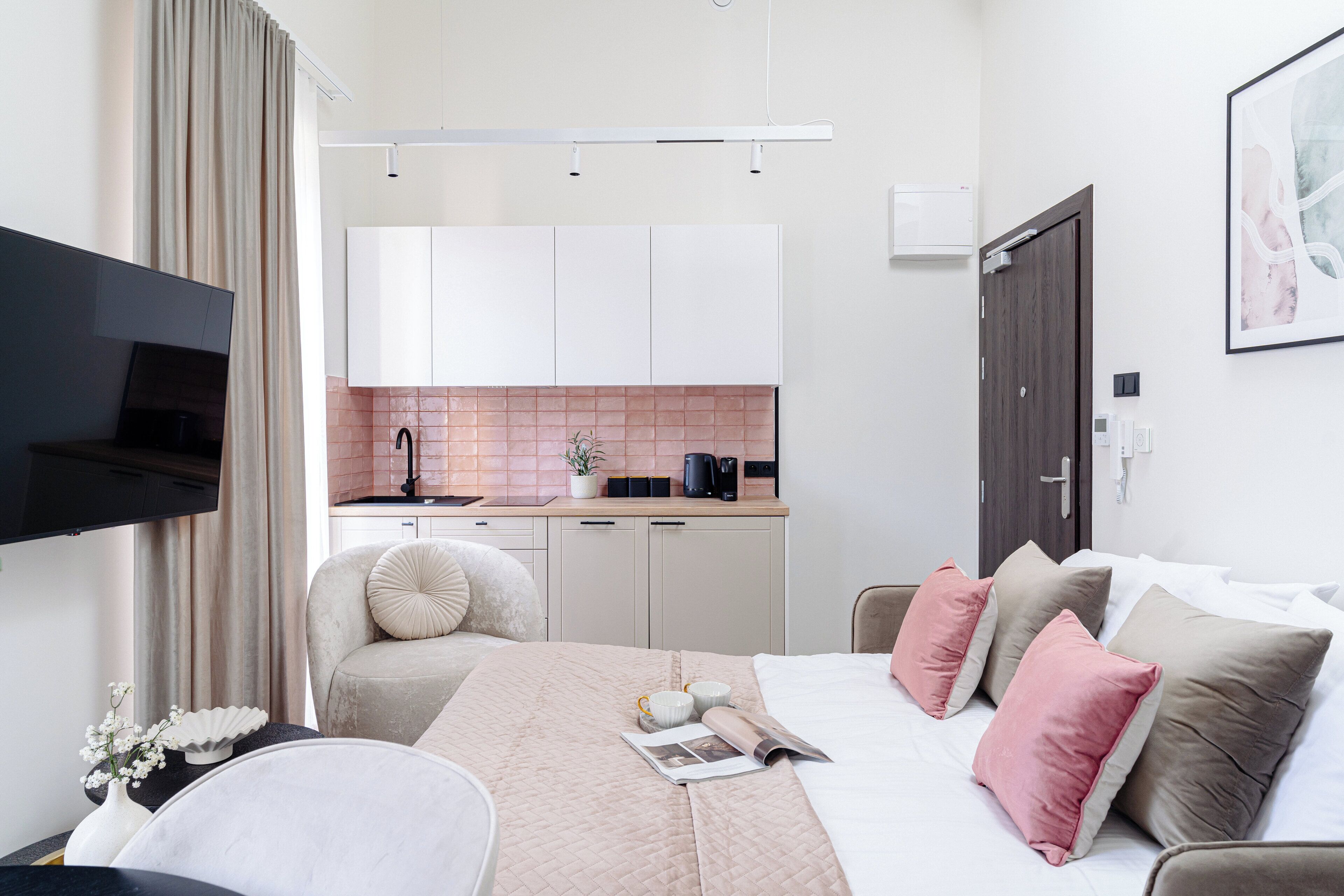Una habitación de hotel con una cama, una mesita de noche con un florero, un televisor, una kitchenette con un salpicadero rosa y una puerta al baño.