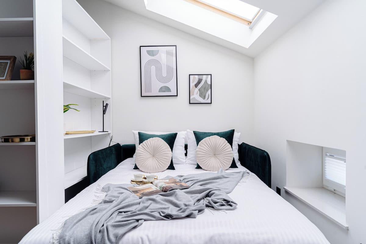 Un dormitorio con tragaluz, paredes blancas, cama con manta gris, dos almohadones decorativos y una mesita de noche con lámpara.