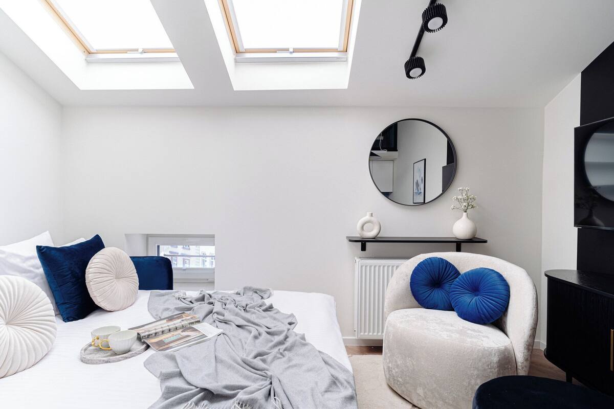 Un dormitorio con tragaluz, un espejo redondo, una cama con almohadas, una silla, un radiador y un televisor.