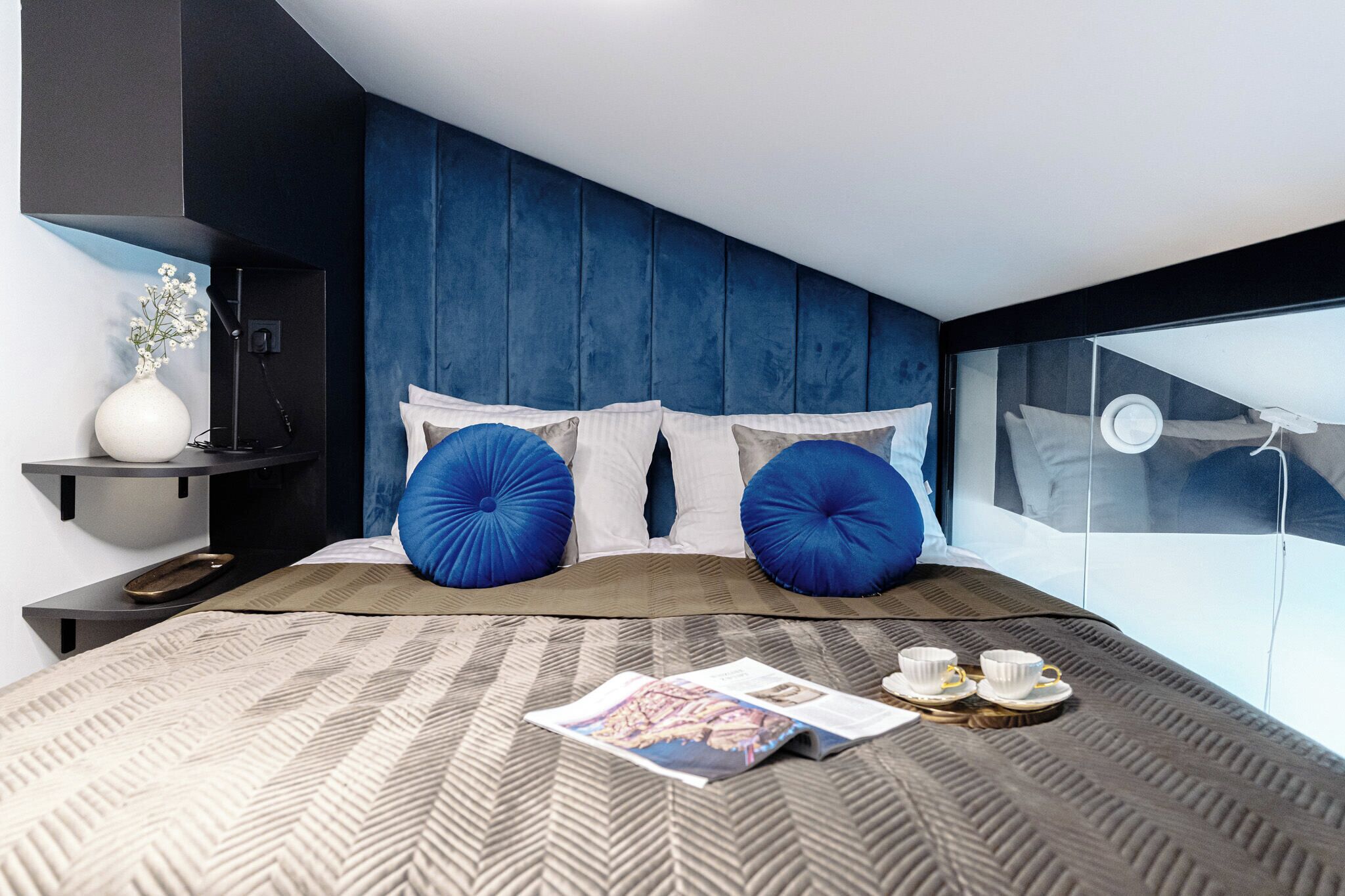 A modern hotel room with a large bed, a bedside table, a magazine, and a tray with cups and saucers.