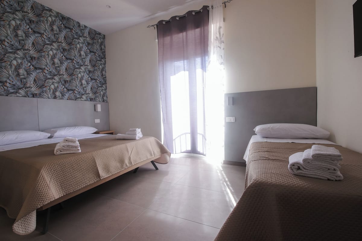 Habitación de hotel con dos camas individuales, un ventanal con cortinas, un papel tapiz estampado y un televisor de pantalla plana montado en la pared.