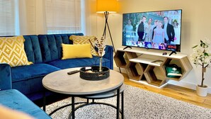 Una sala de estar con un sofá azul, una mesa de centro, un televisor sobre un soporte de madera, una lámpara y un reloj en la pared.