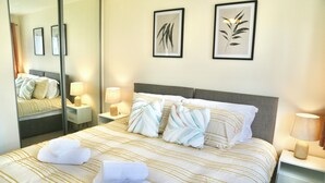 Una cama bien tendida con ropa de cama a rayas y cojines decorativos, una mesita de noche con lámpara, y dos cuadros enmarcados en la pared.