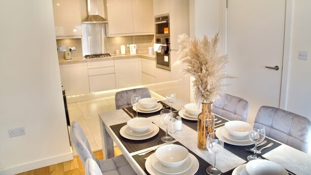 Un comedor moderno con una mesa dispuesta para cuatro, un jarrón con plumas de pampa y una cocina al fondo.