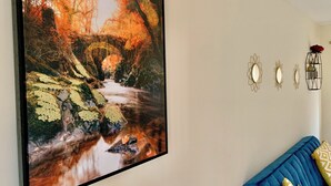 Una habitación con un cuadro grande enmarco de una escena de bosque, un sofá azul con cojines estampados, y tres espejos decorativos colgados en la pared.