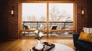 A spacious room with a large sliding glass door leading to an outdoor patio. There is a wooden floor, a white vase with flowers, and a dark-colored sofa with a yellow cushion. - Brick & Balcony Greeley Retreat (Greeley)