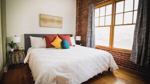 A bedroom with a large bed, a brick wall, a window with curtains, and a framed picture on the wall. - Brick & Balcony Greeley Retreat (Greeley)