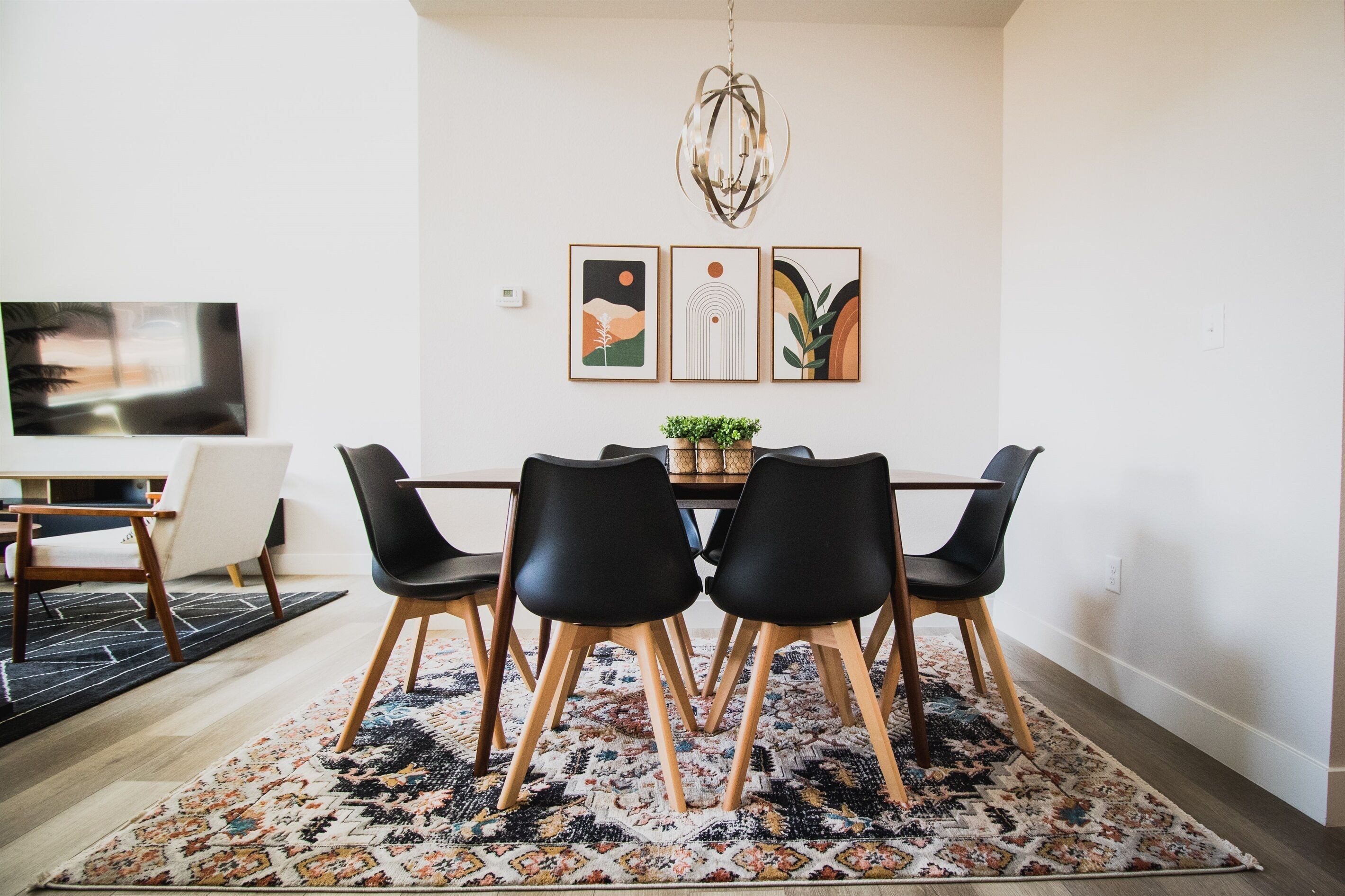 A dining area with a wooden table, black chairs, and a patterned rug. There are three framed artworks on the wall above the table.
