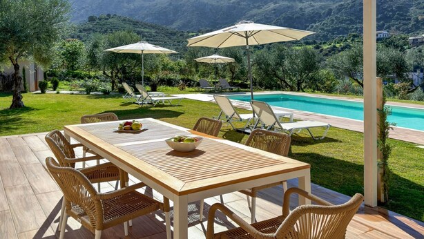 A poolside area with a wooden table and wicker chairs, surrounded by umbrellas and a view of mountains. - Villa Adam (Chania)