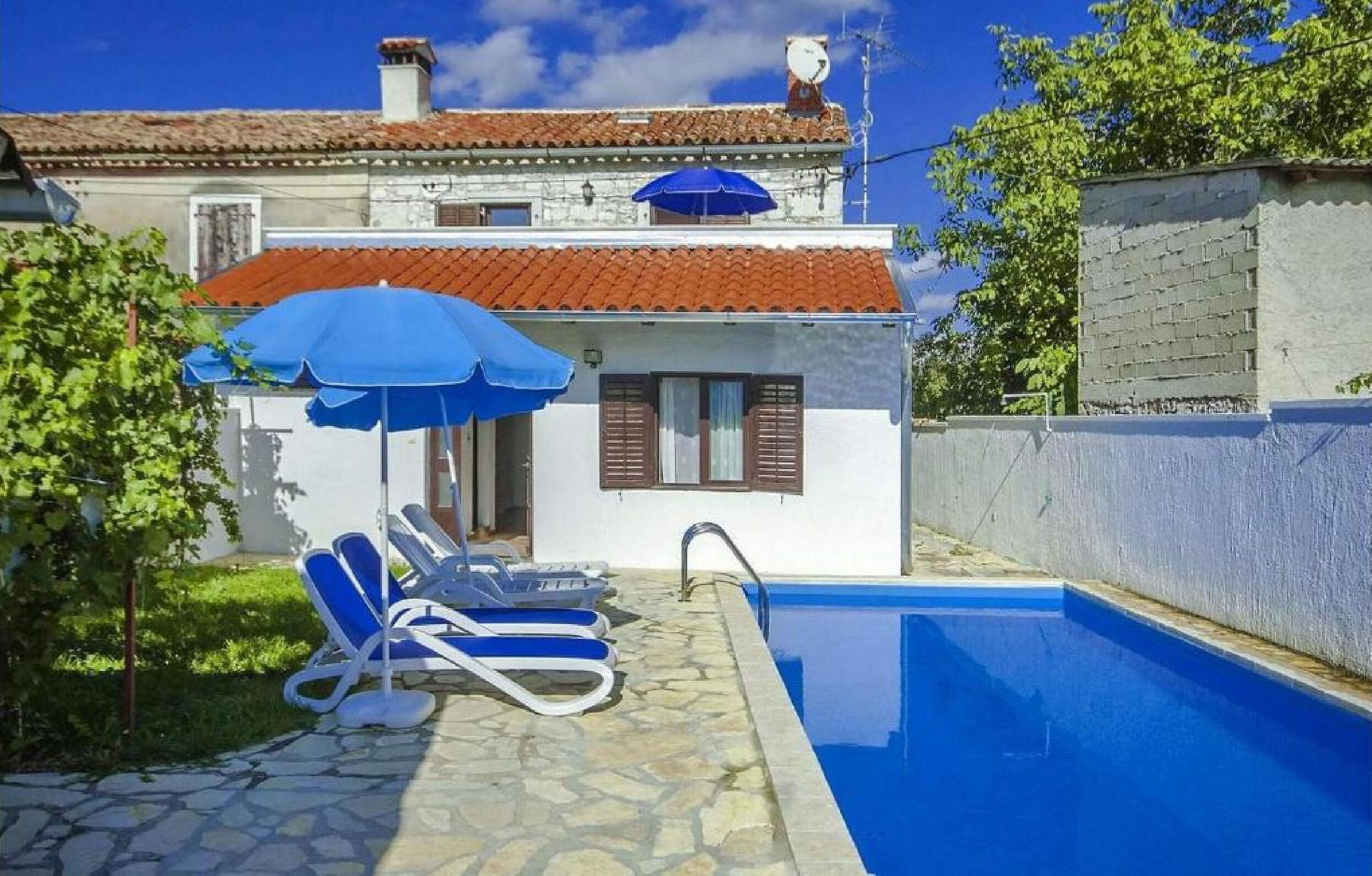 A pool area with lounge chairs and a blue umbrella, a white building with a red-tiled roof, and a stone pathway.