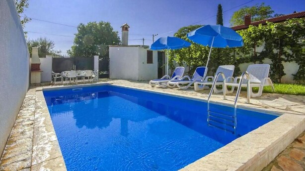 Una piscina rettangolare con superficie d'acqua azzurra e trasparente, circondata da lettini bianchi e ombrelloni blu.