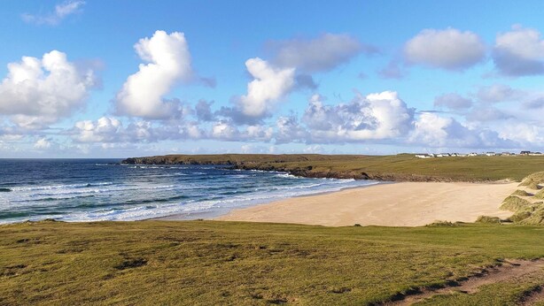 Paisagem costeira com uma praia arenosa, áreas gramadas e vista para o oceano.