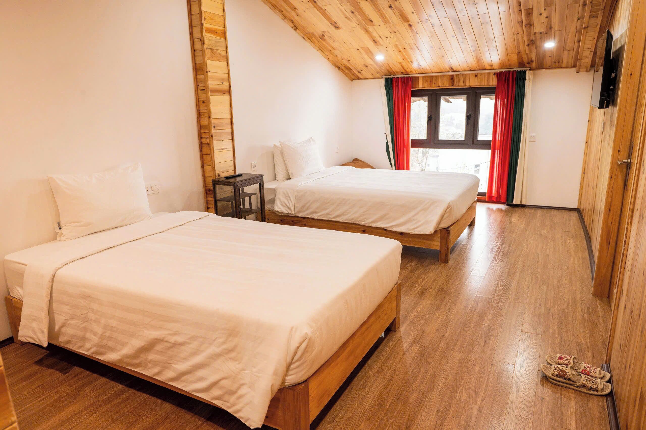 A room with two beds, wooden flooring, a wooden ceiling, and a window with red curtains.