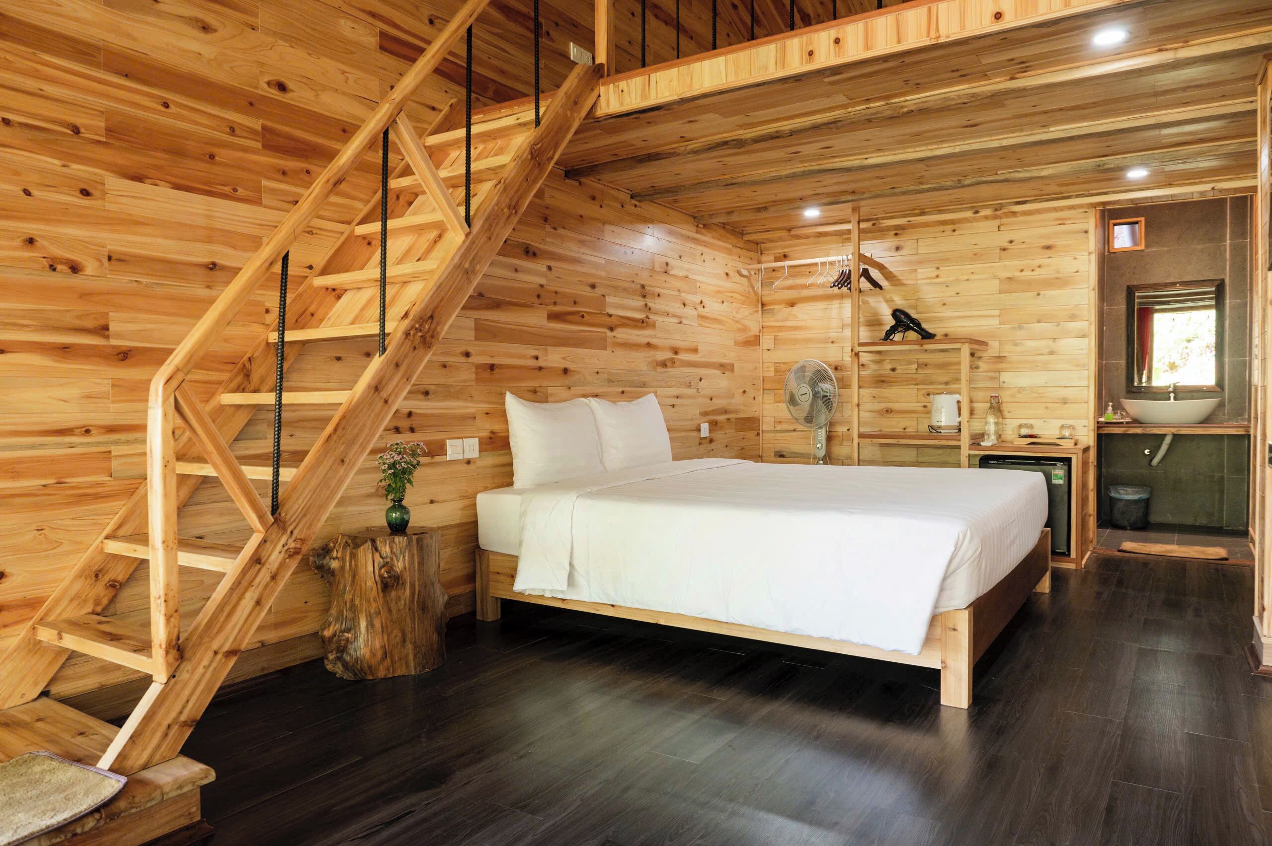 Un dormitorio de cabaña de madera con una cama, una escalera de madera, una mesita con un ventilador y una ventana con vistas.