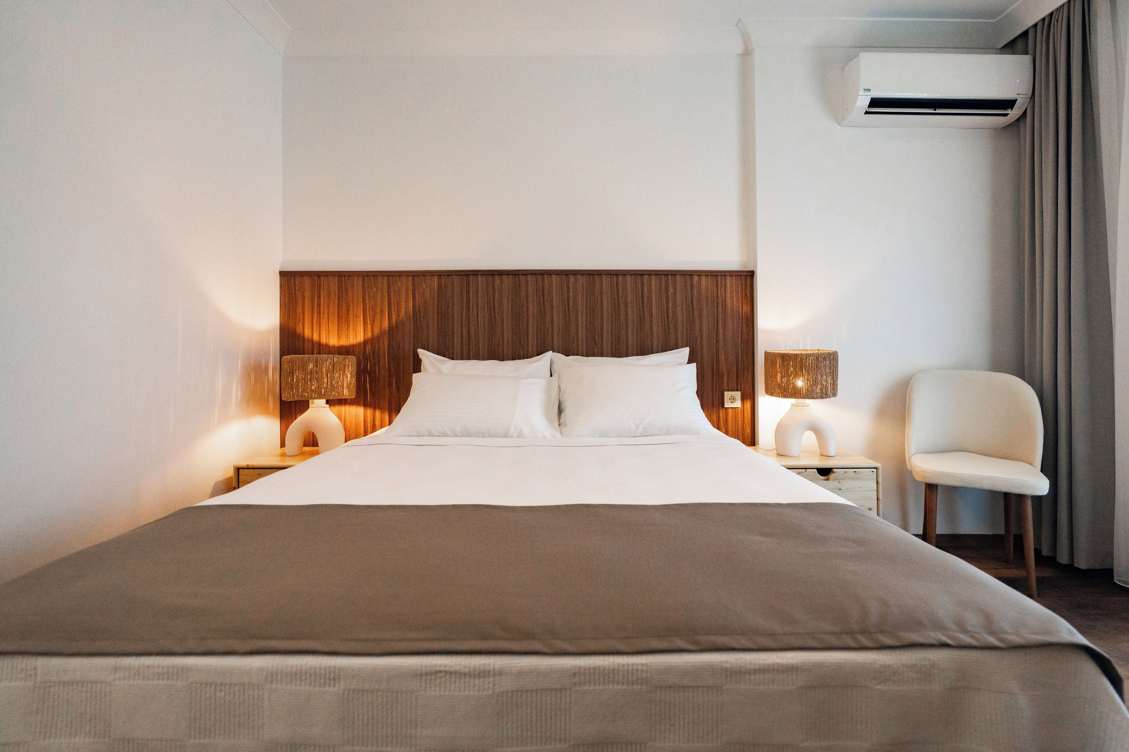 A hotel room with a large bed, two bedside lamps, a chair, and an air conditioning unit.