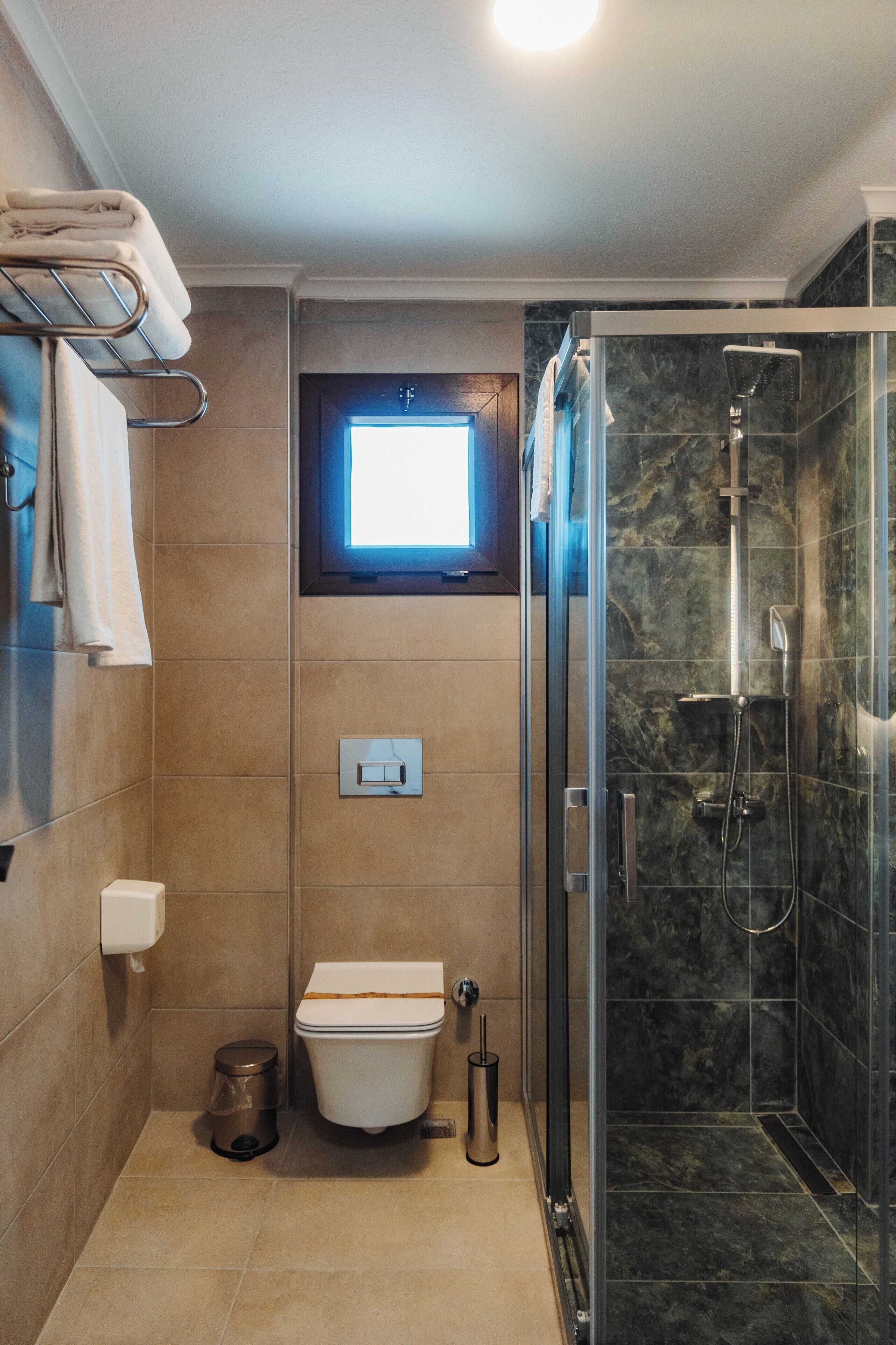 A bathroom with a toilet, a trash can, a towel rack, a window, and a glass-enclosed shower.