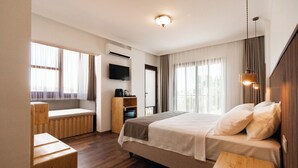 Quarto de hotel com uma cama grande, um banco de madeira, uma TV e uma janela com cortinas.