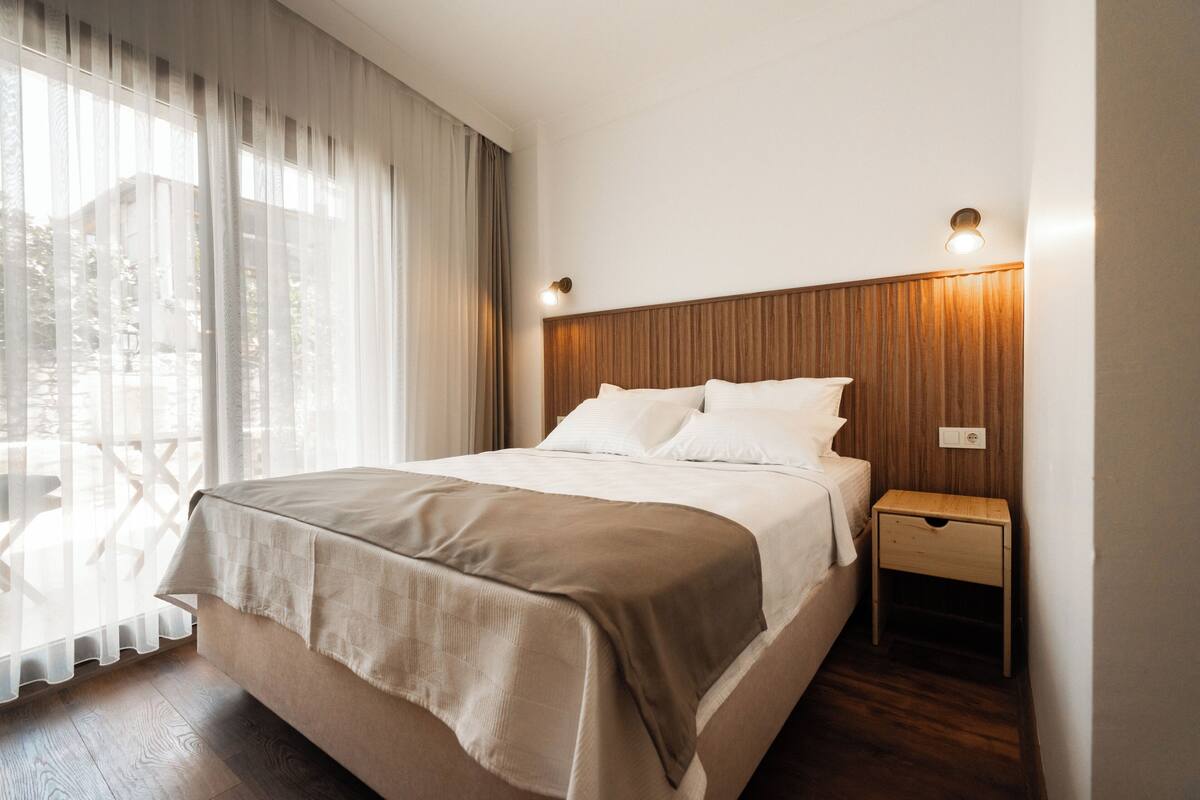 A hotel room with a bed, a nightstand, a wooden headboard, and a view of the outdoors through sheer curtains.