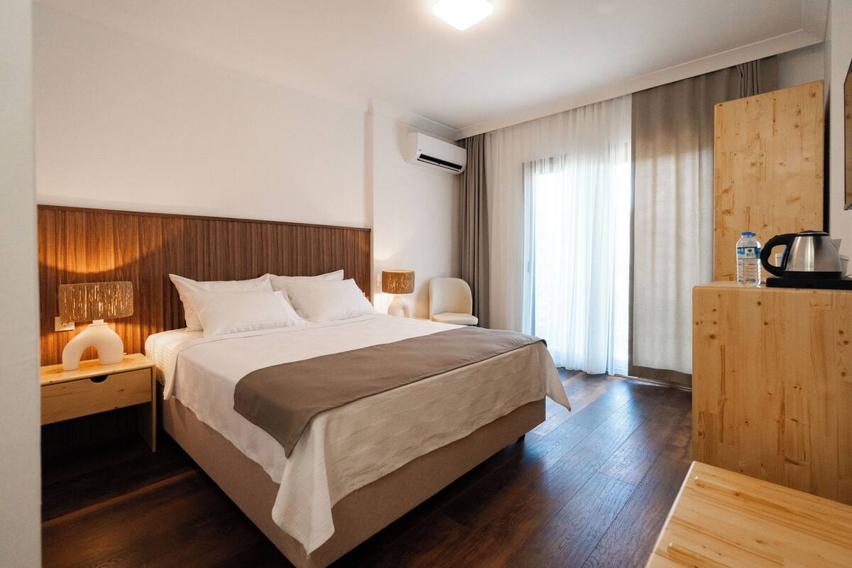 A hotel room with a large bed, a wooden headboard, a nightstand with a lamp, a wooden dresser, and an air conditioning unit.