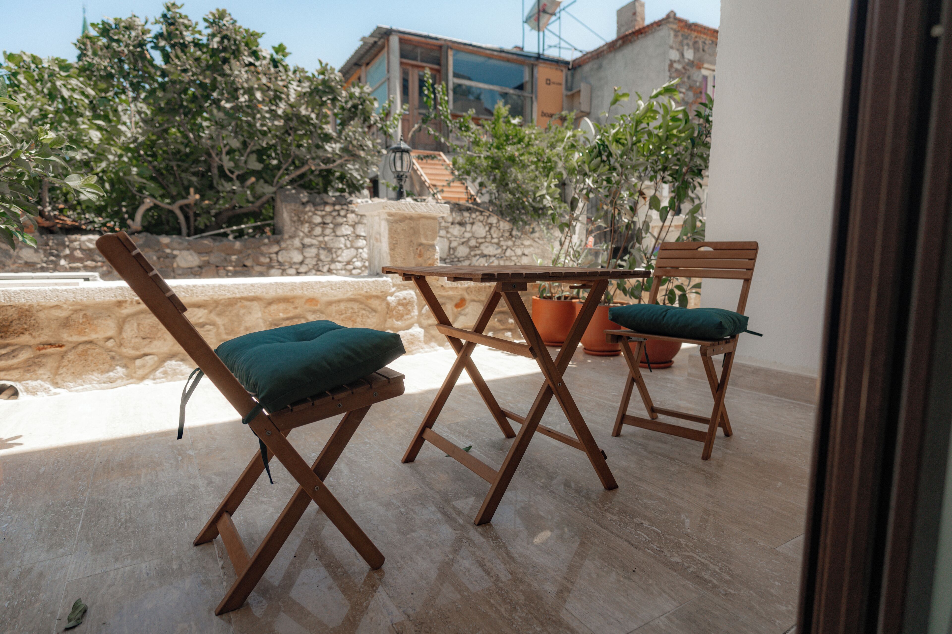Un petit espace extérieur aménagé avec des chaises et une table en bois, entouré de plantes en pot et d’un mur de pierres.
