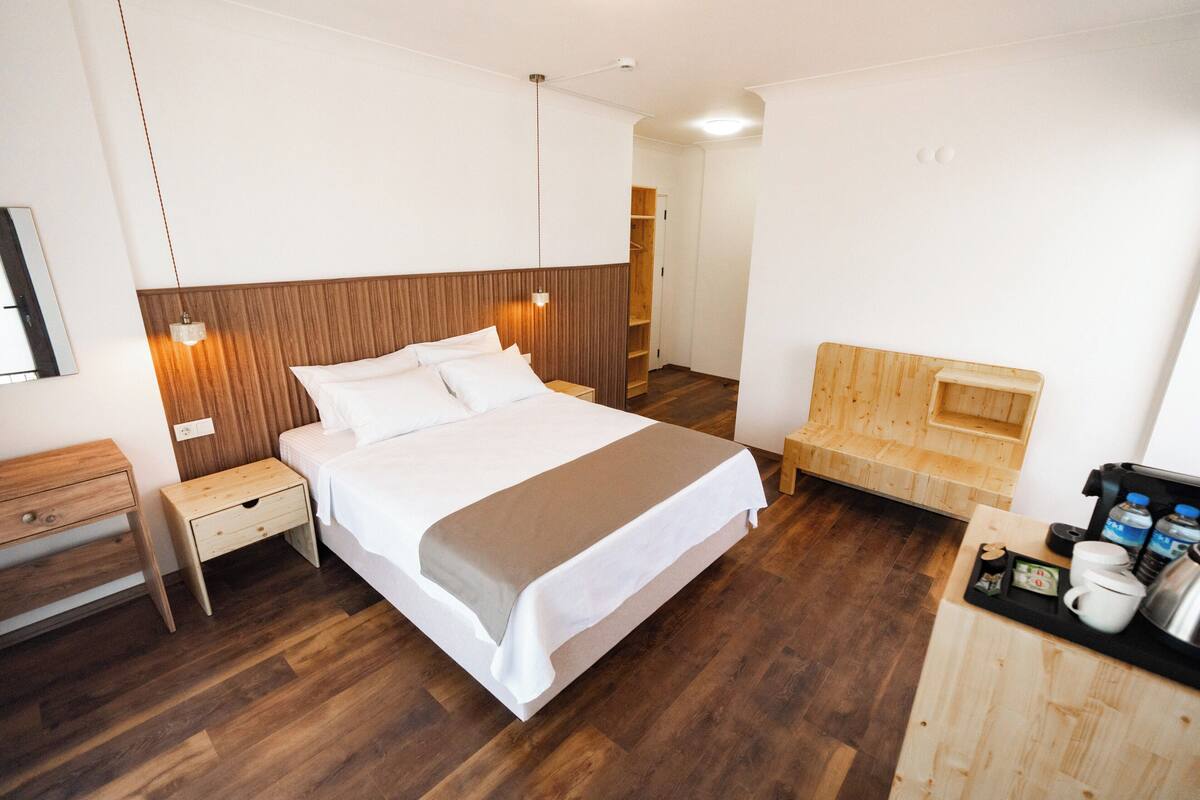 A modern hotel room with a wooden headboard bed, a wooden nightstand, a wooden bench, and a small kitchenette area with a coffee maker and water bottles.