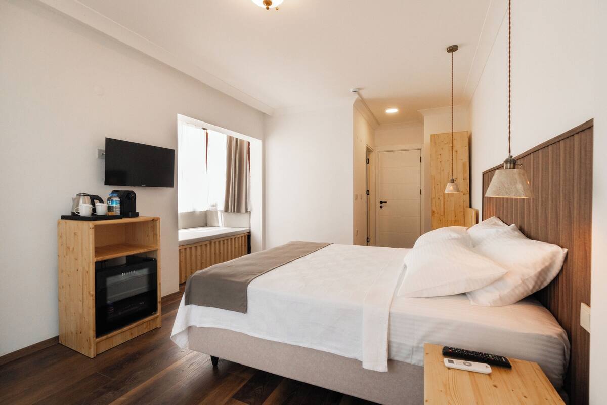 A modern hotel room with a bed, a TV, a wooden shelf with a coffee maker, and a wooden headboard.