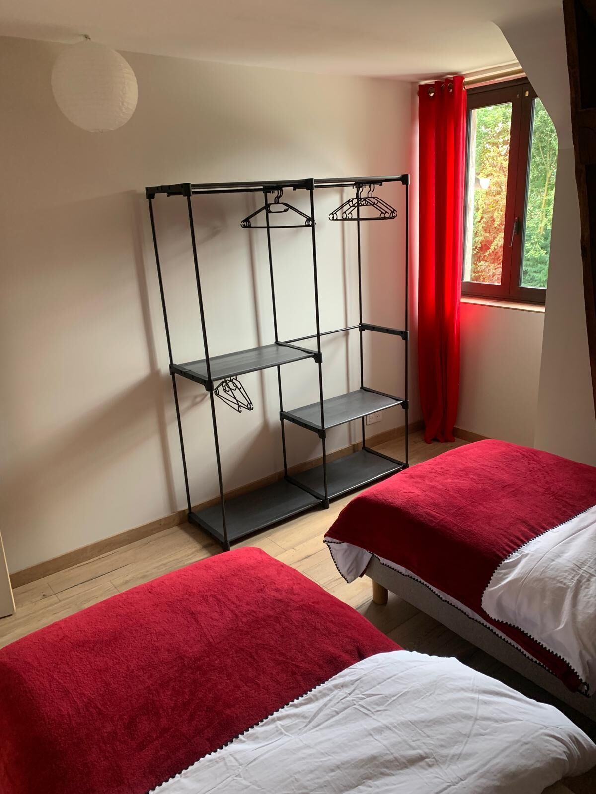 Quarto com duas camas, um guarda-roupa de metal, uma janela com cortinas vermelhas e uma luminária de teto branca.