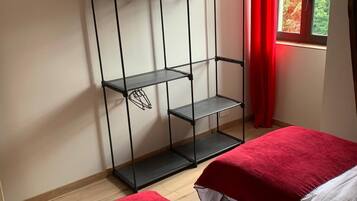 Quarto com duas camas, um guarda-roupa de metal, uma janela com cortinas vermelhas e uma luminária de teto branca.