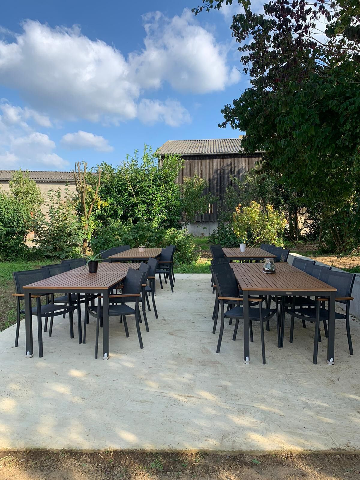 En udendørs terrasse med flere træborde og sorte stole, omgivet af grøn vegetation og en bygning i baggrunden.