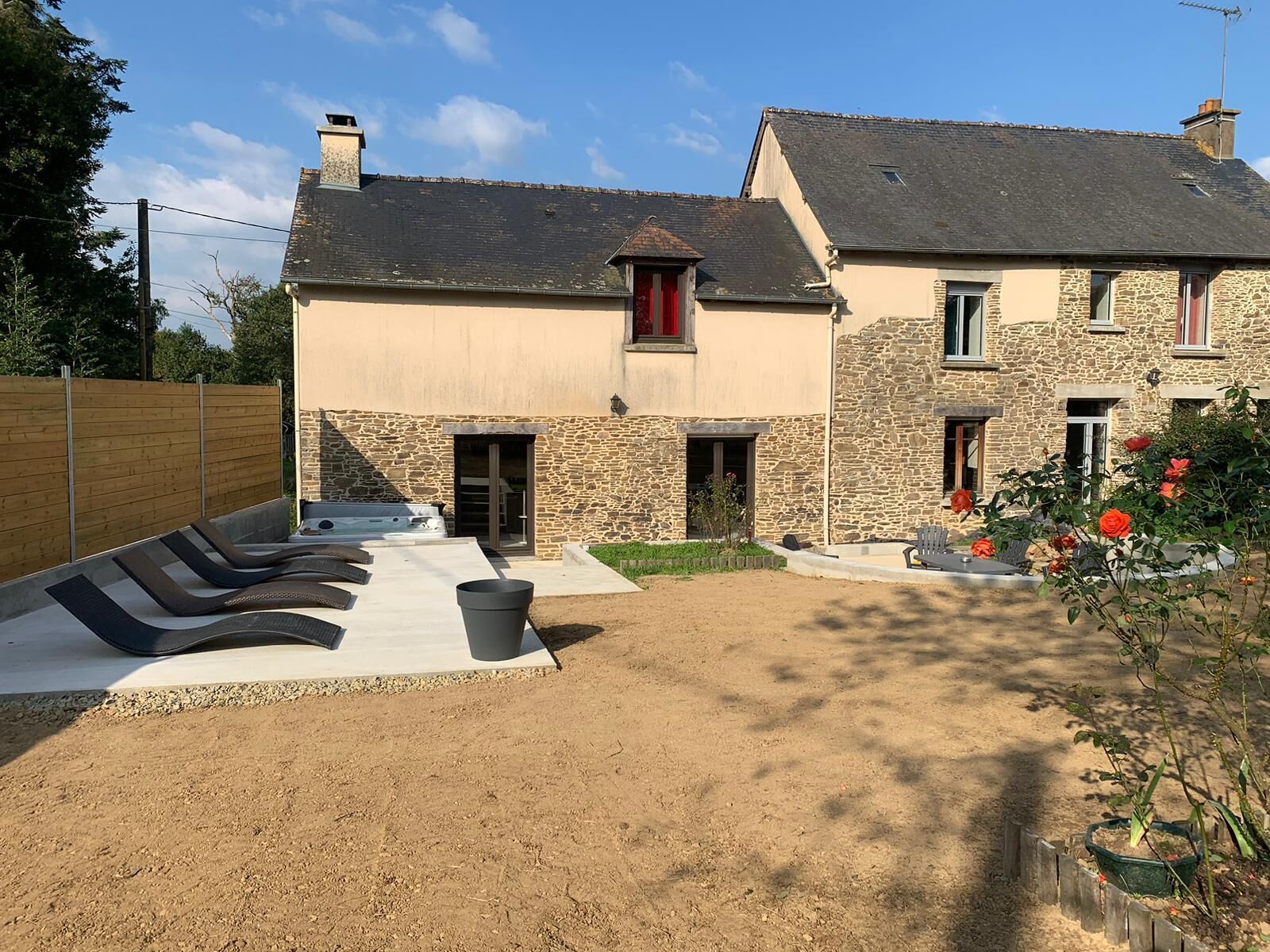 Une maison en pierre à deux étages, avec une façade en pierre, un petit jardin fleuri et une terrasse équipée de chaises longues.