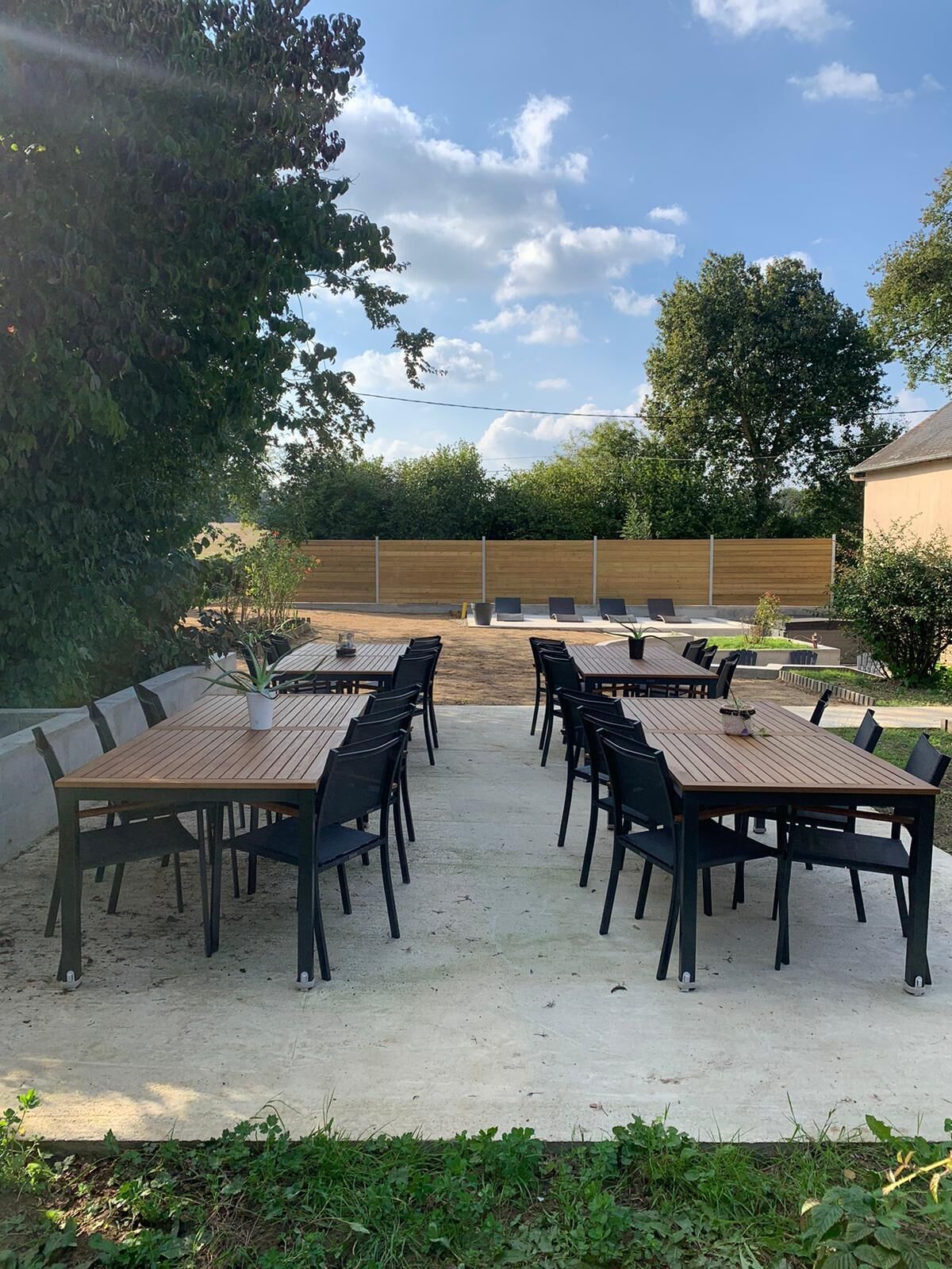 An outdoor patio with multiple wooden tables and chairs, surrounded by a low wooden fence and greenery.