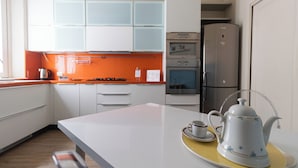 Una cocina moderna con armarios blancos, salpicadero naranja, electrodomésticos de acero inoxidable y encimera blanca con tetera y taza.