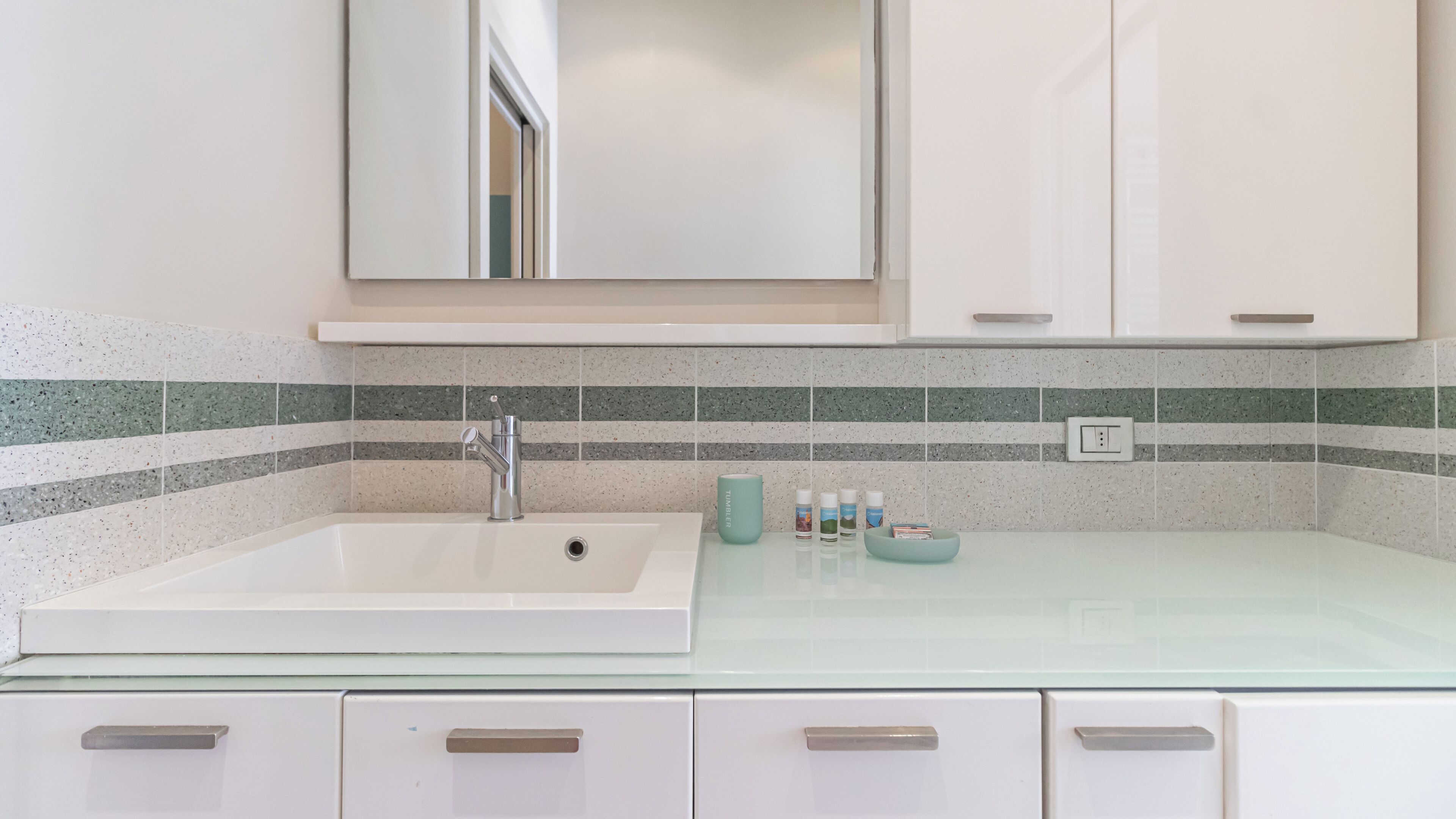Een badkamer met een witte wastafel, een spiegel erboven en een betegelde spatwand. Onder de wastafel bevinden zich kastjes en een aanrecht met toiletartikelen.