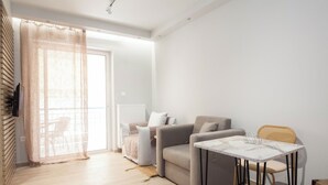 A modern living room with a sofa, a dining table, and a chair. There is a large window with a sheer curtain, a radiator, and a wicker chair. - Petralona Gem Hotel-Style Studio Balcony (Athens)