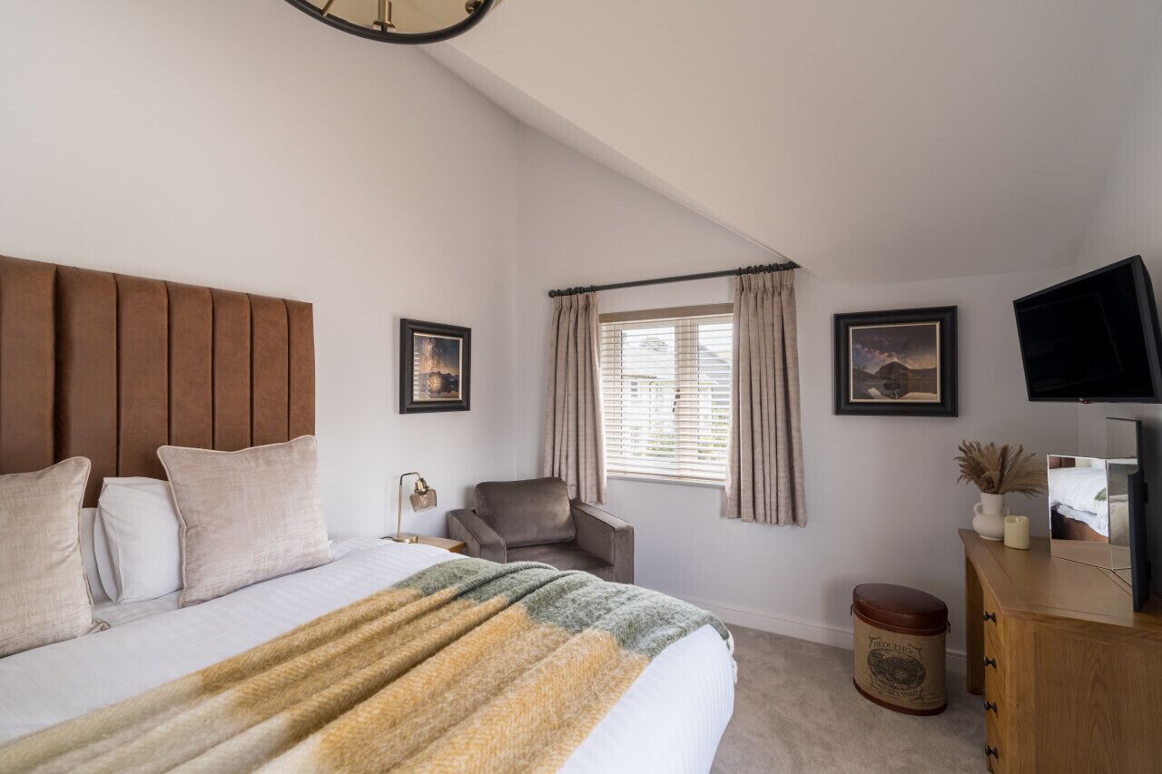 Un dormitorio con una cama grande, cabecera de madera, una silla, televisión y ventana con cortinas.