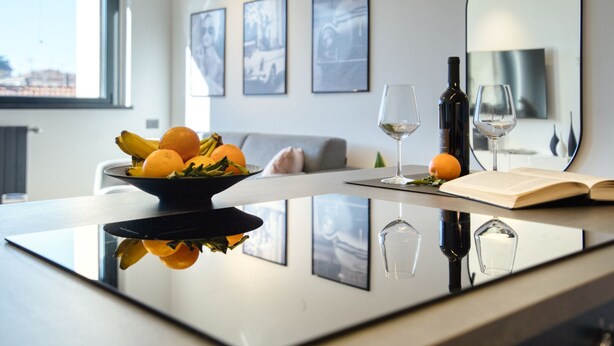 A modern kitchen with a glass-top island, a bowl of fruit, wine glasses, and a book. - Canal Couture Navigli Luxury Retreat (Milan)