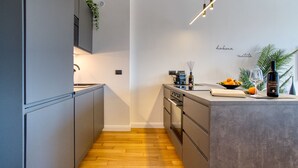 A modern kitchen with grey cabinetry, a wooden floor, and a built-in oven. There is a small island with a wine bottle and fruit on it. - Canal Couture Navigli Luxury Retreat (Milan)