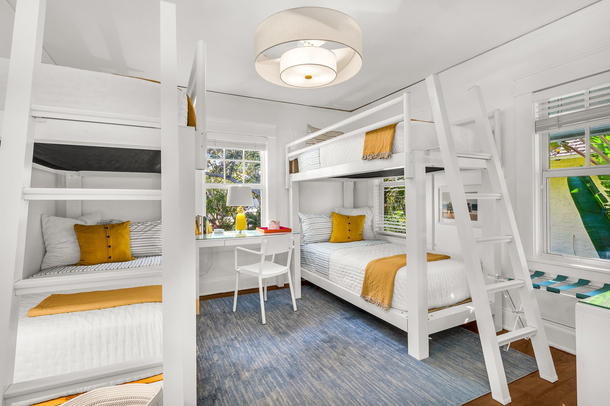 A bunk bed room with a desk and ladder, white walls, and a window with a view.