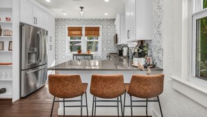 A modern kitchen with white cabinets, a stainless steel refrigerator, a central island with bar stools, and a window with blinds. - Casa Mía - Palm Beach Boho Escape (West Palm Beach)